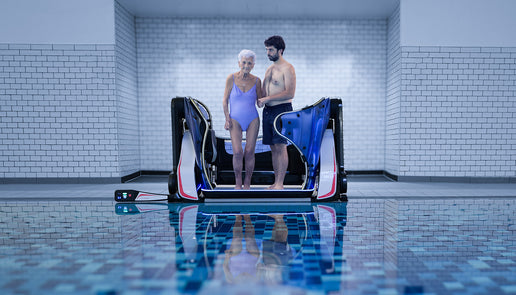 A striking image of a pool lift from the front. The image shows an elderly woman wearing a swimming costume being assisted by a man. The background is white tiles with black grout, the foregrount is is blue tones of swimming pool water. The image of the striking and elegant pool lift is reflected in the water in the foreground making a beautiful composition.